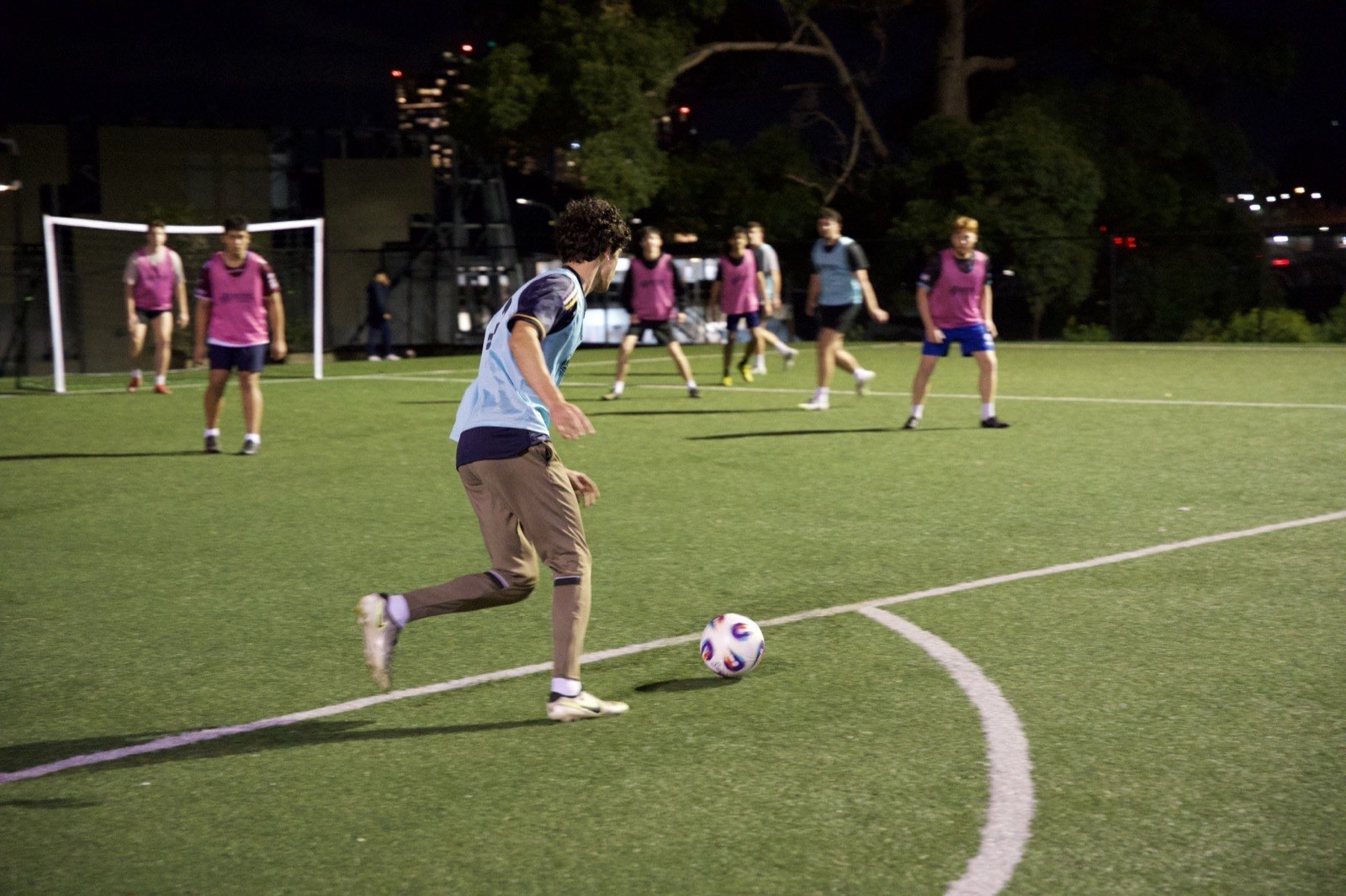 A curly-haired player in a light-blue bib dribbling the ball forward on a floodlit pitch