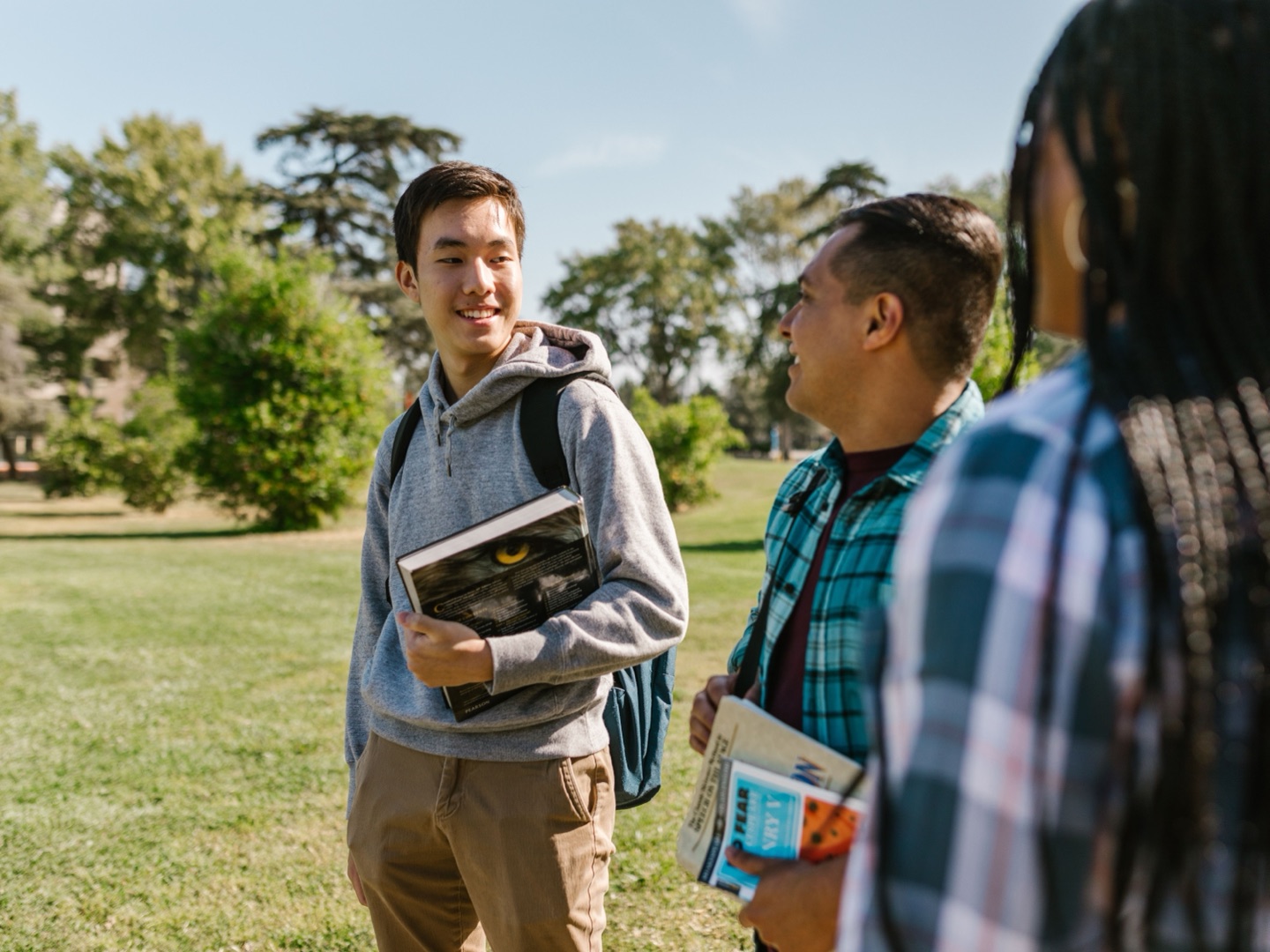 Three students chatting on a campus lawn, one holding a textbook